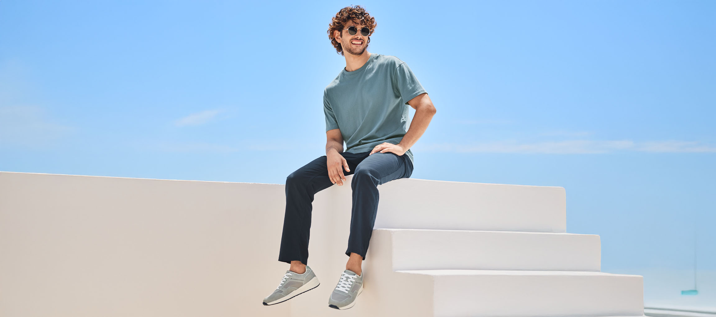 Мъж в тениска, панталон и маратонки New Balance, седнал на бяло стълбище.