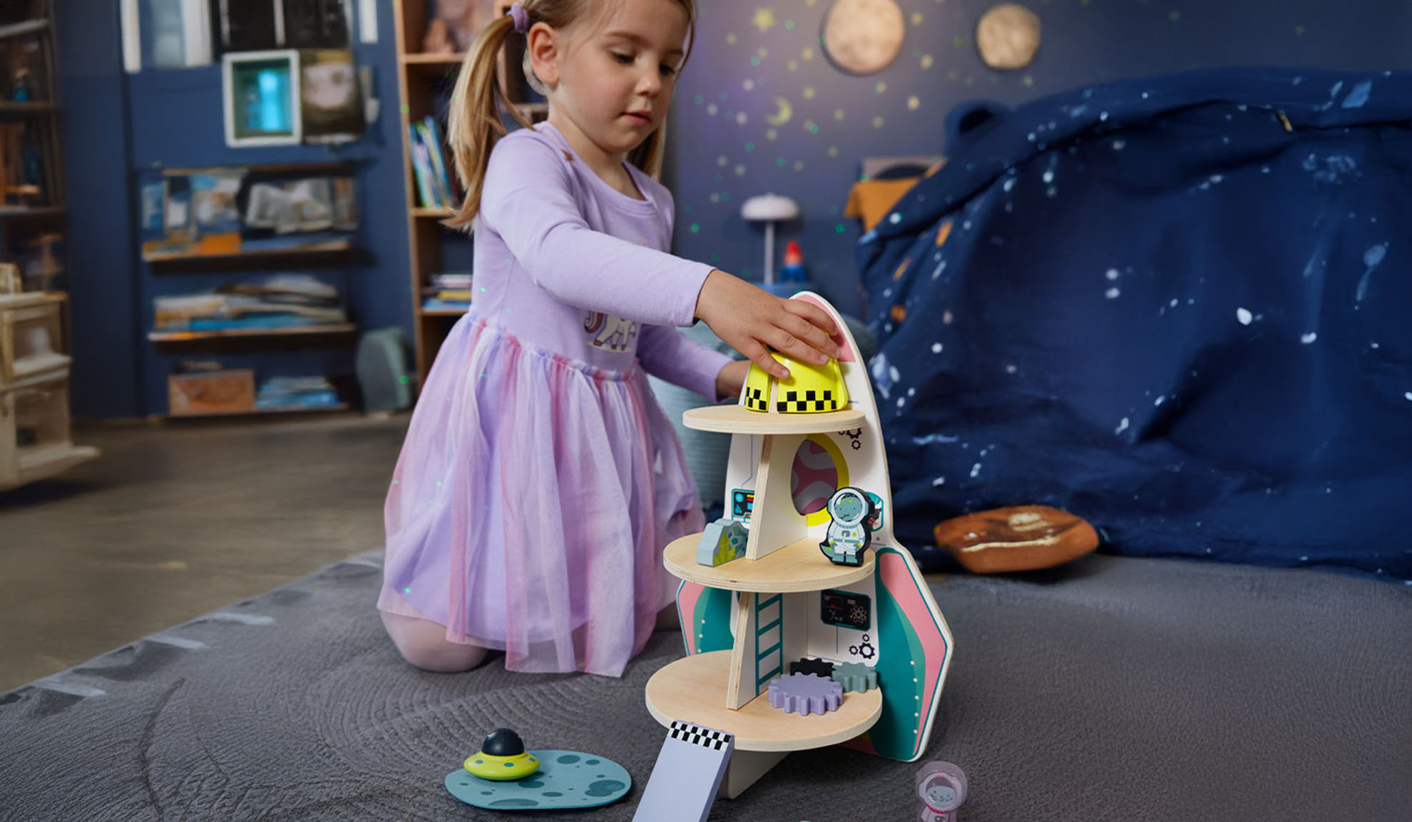 Момиче си играе с играчка ракета и космически аксесоари в тематична стая.