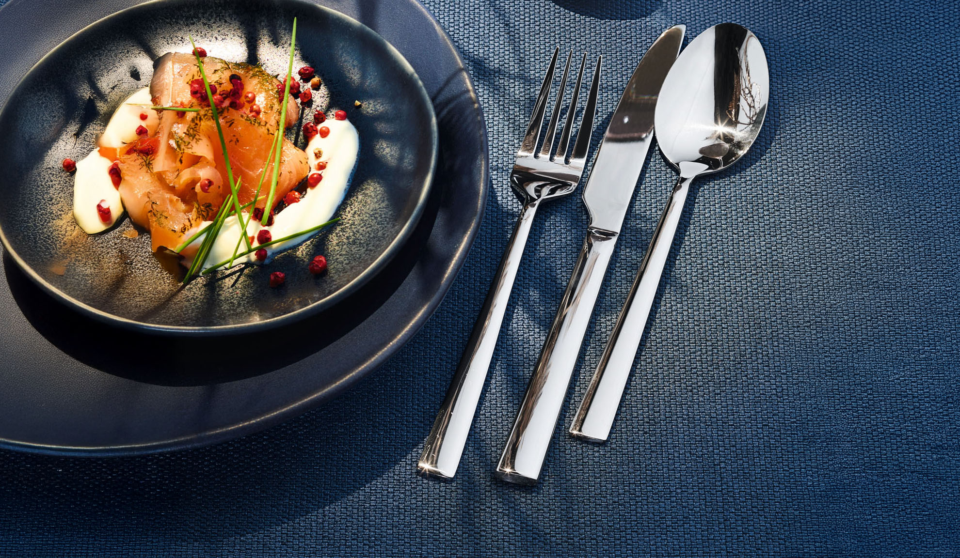 Пушена сьомга със сметана, билки и розов пипер върху тъмна чиния с прибори.