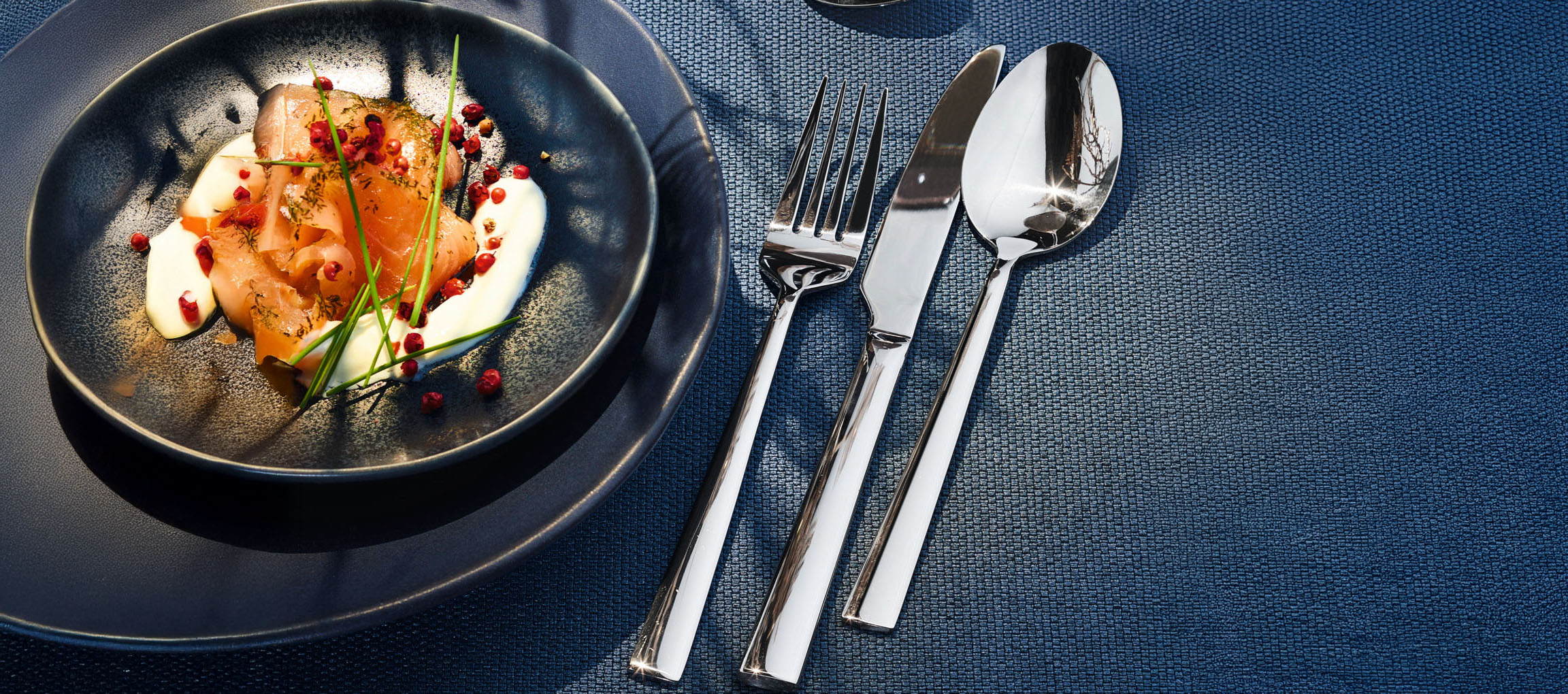 Пушена сьомга със сметана, див лук и розов пипер върху тъмна чиния с прибори.