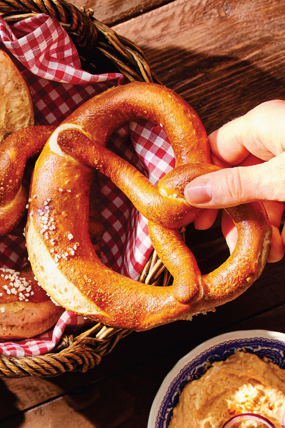 Ръка взима солен геврек от кошница с червено-бяла карирана кърпа, до купа с дип.