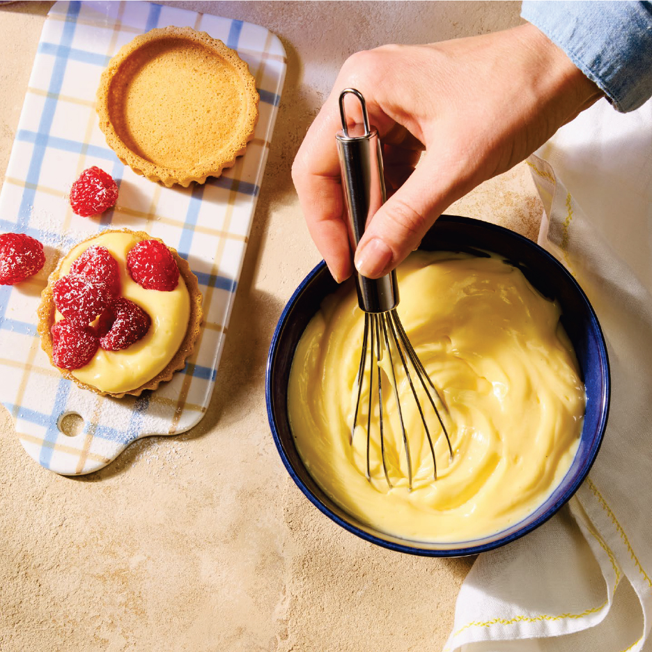 Ръка разбърква крем в купа, докато на дъска има тарталети с малини.