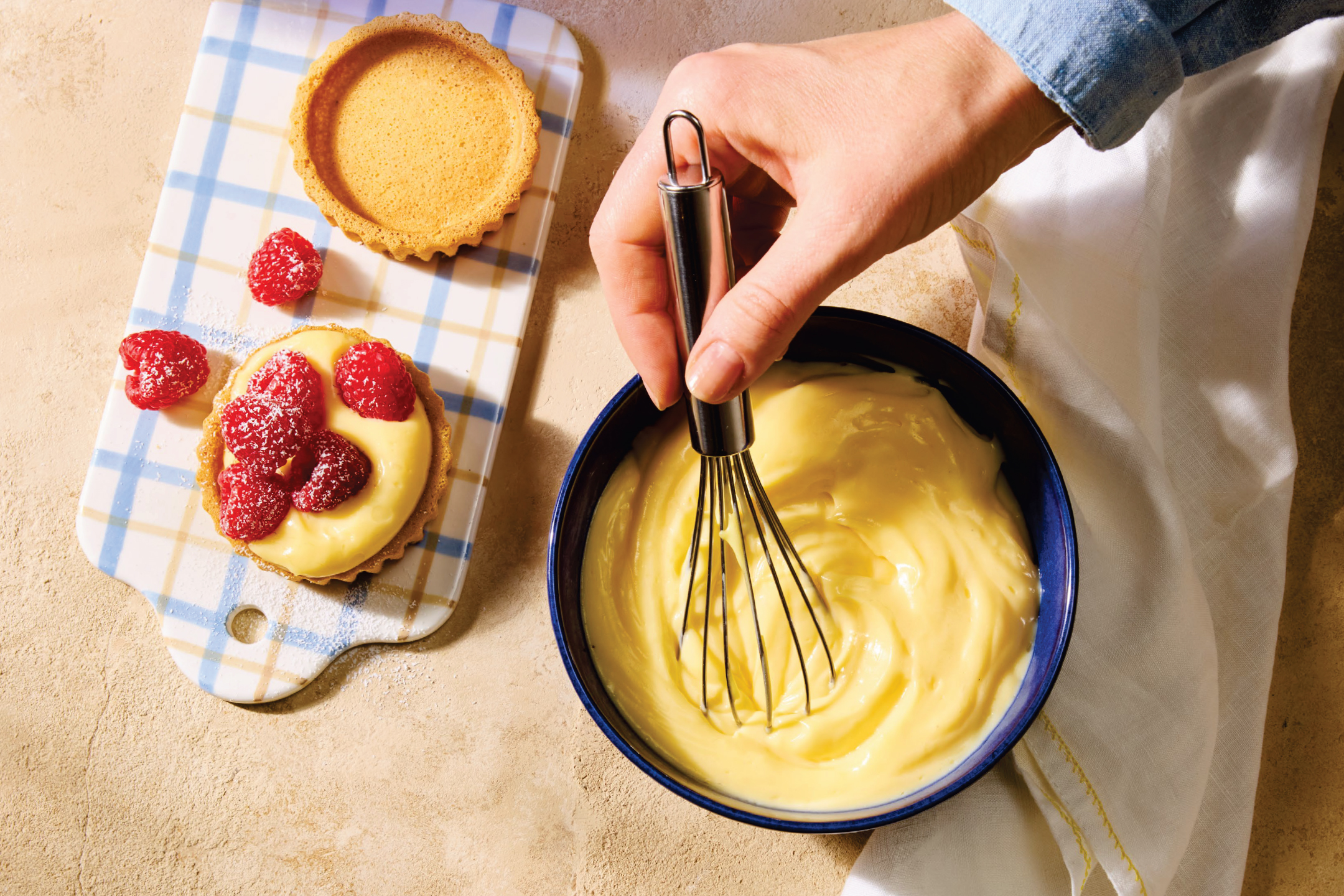 Ръка бърка крем в купа, докато на дъска има тарталети с малини и пудра захар.