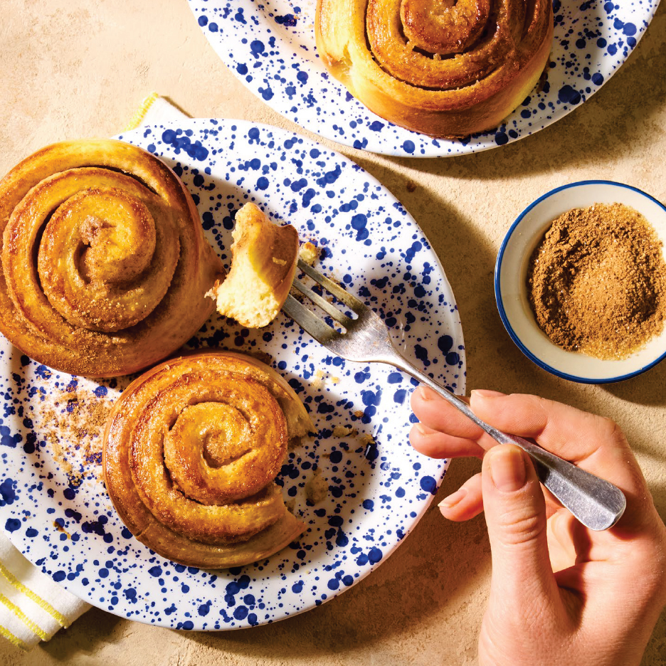 Канелени рулца върху сини и бели чинии, с вилица, която взима парче, и купа с канела.