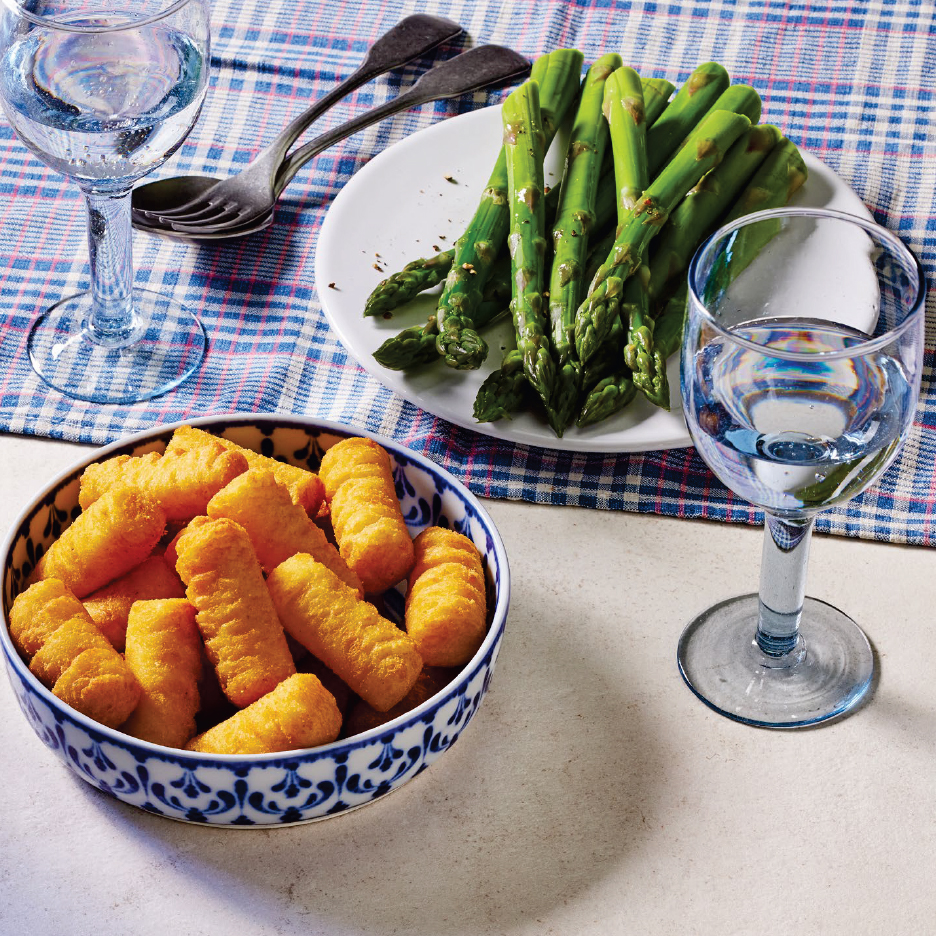 Купа с картофени крокети, чиния със зелени аспержи и чаши с вода на карирана покривка.