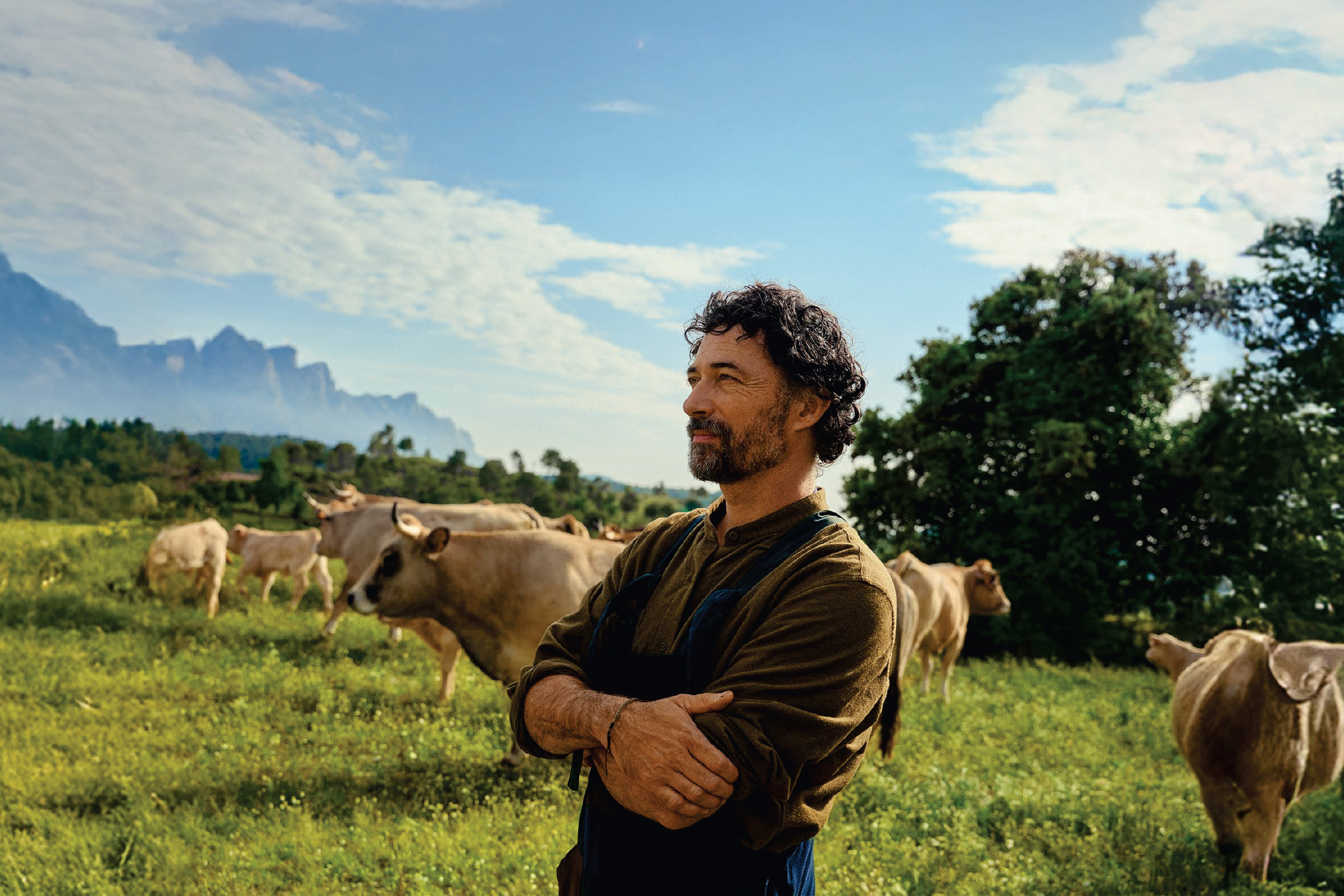 Фермер с брада, облечен в кафява риза и сини гащеризони, стои сред крави на пасище.