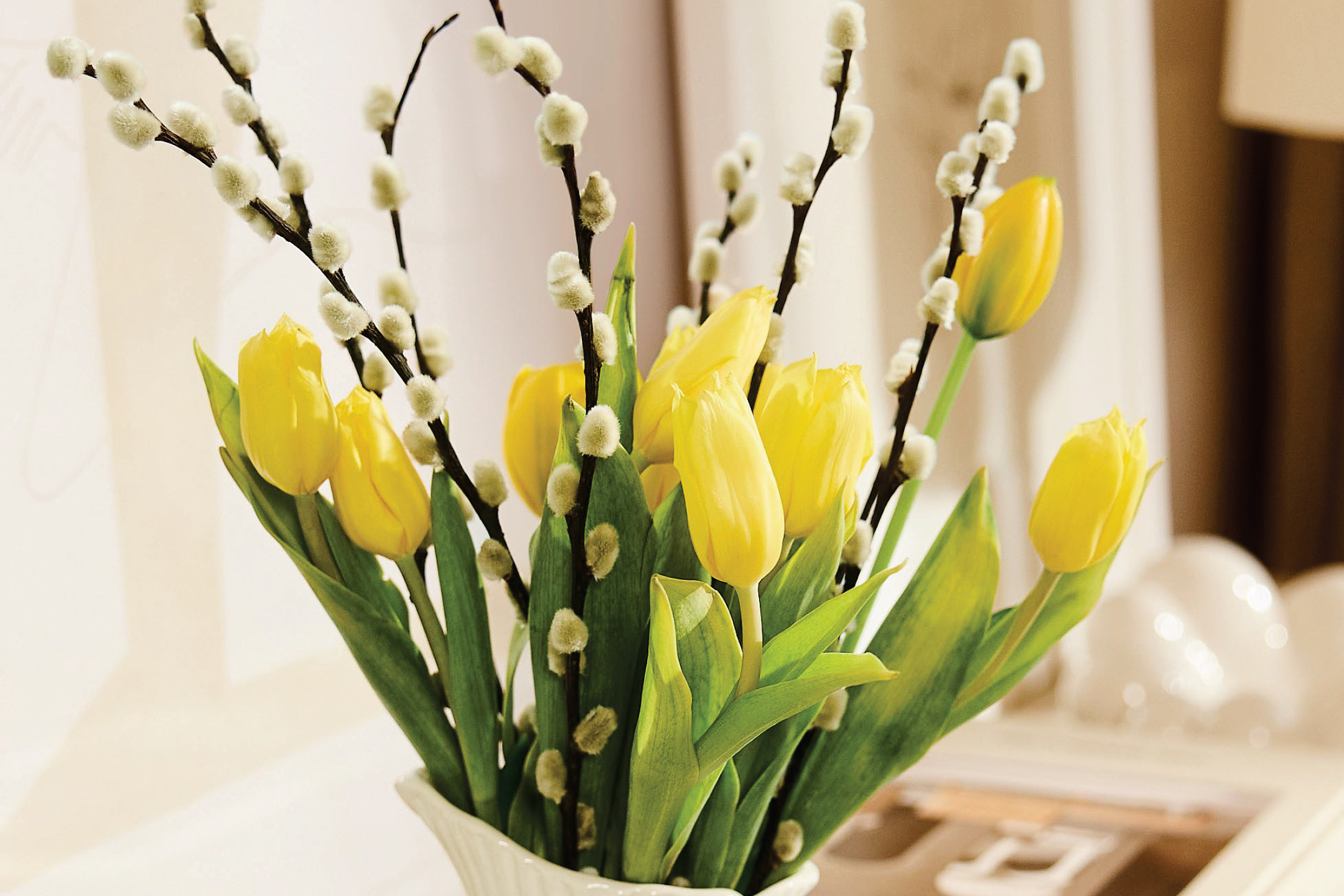 Букет от жълти лалета и върбови клонки в бяла ваза, създаващ великденска атмосфера.