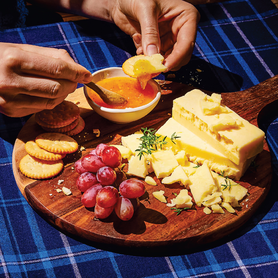 Дъска със сирене, крекери, грозде и сладко, с ръце, потапящи крекер в сладко.