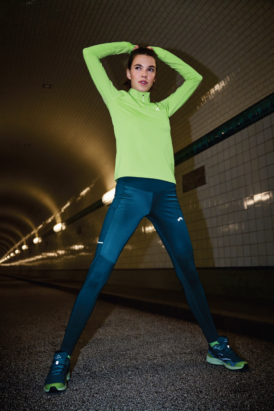 Жена в спортен екип Crivit: зелена блуза с дълъг ръкав, тъмносини клинове и маратонки.