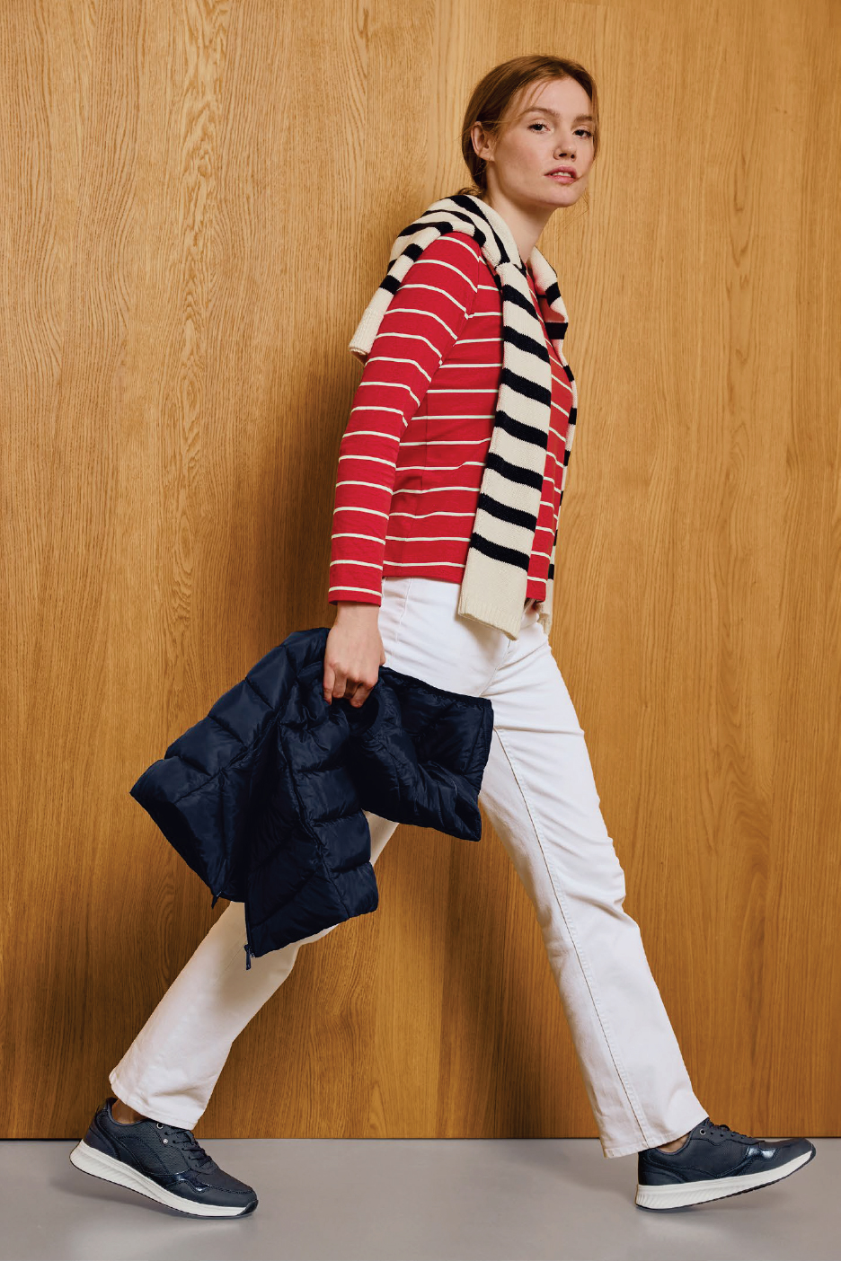 Жена в червена раирана блуза, бели панталони и тъмносини маратонки, носеща тъмносиньо яке.