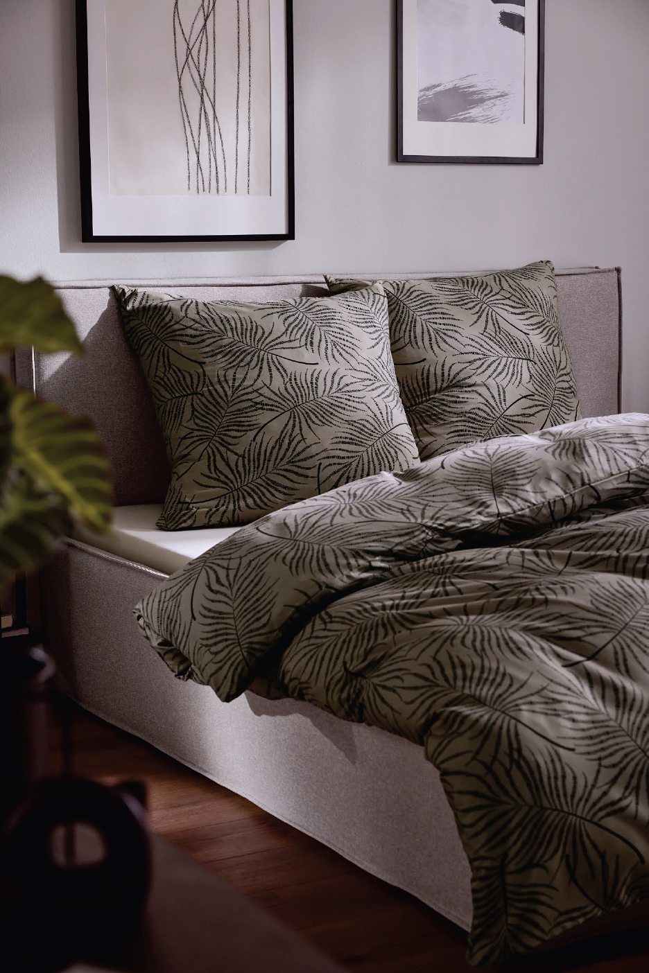 Спалня с бежово спално бельо с щампа на палмови листа и две картини на стената.