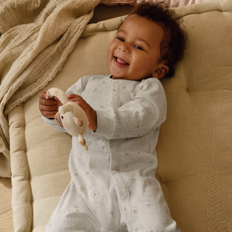 Усмихнато бебе в гащеризон, държащо плетена играчка, лежи на бежова възглавница.