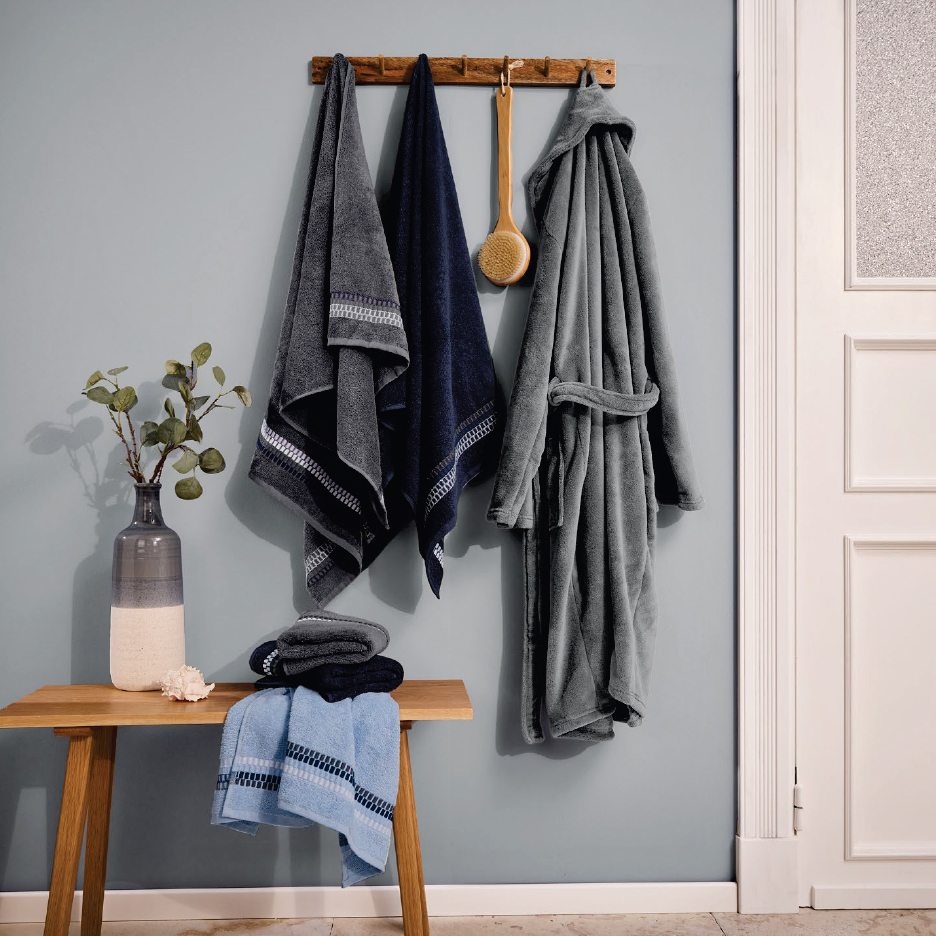 Сиви и сини хавлиени кърпи и халат за баня, висящи на стена, с пейка и ваза.
