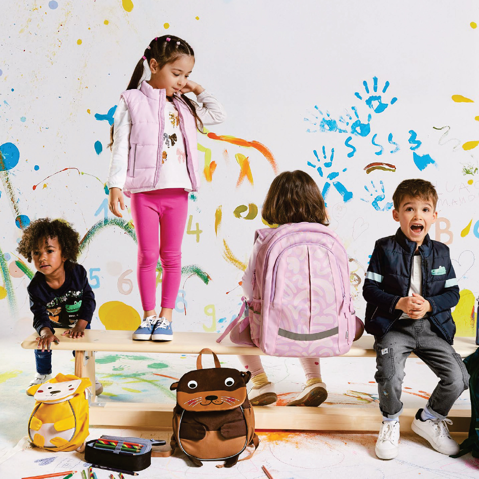 Деца с раници и аксесоари за училище пред изрисувана стена.