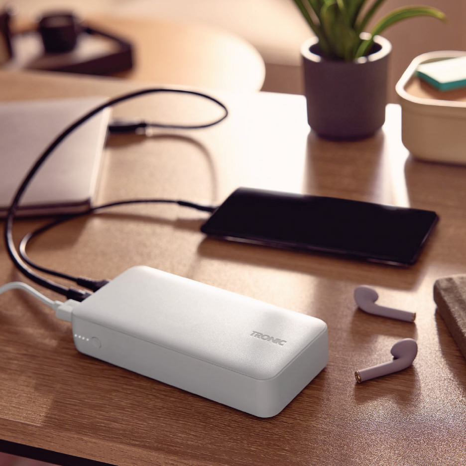 Бяла външна батерия, зареждаща смартфон и лаптоп на дървена маса, с безжични слушалки.