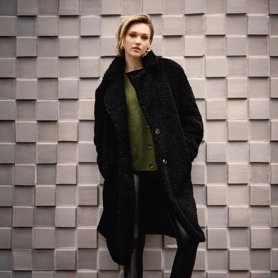 Жена в черно палто от букле, зелена жилетка и черни кожени панталони, позираща до стена.