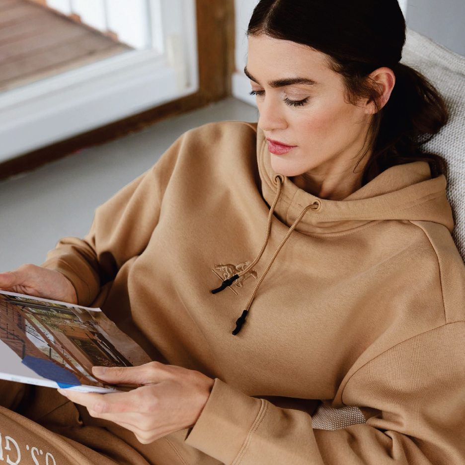 Жена в бежов суичър с качулка чете списание, докато седи на диван.