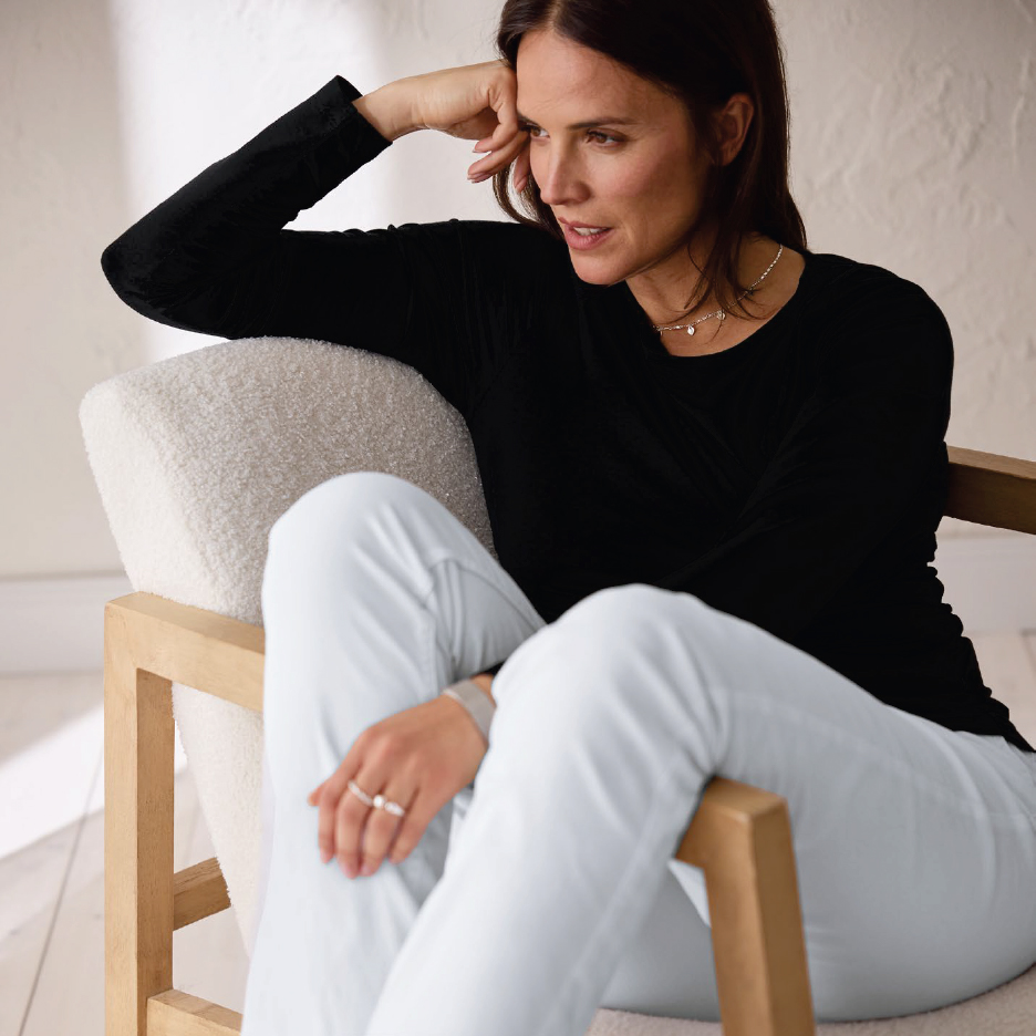 Жена в черен пуловер и светли панталони седи на стол, замислена.