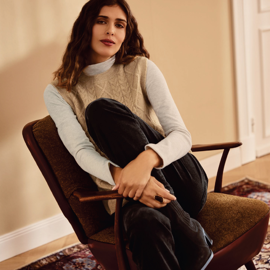 Жена в бежов плетен елек и черни панталони, седнала на стол.