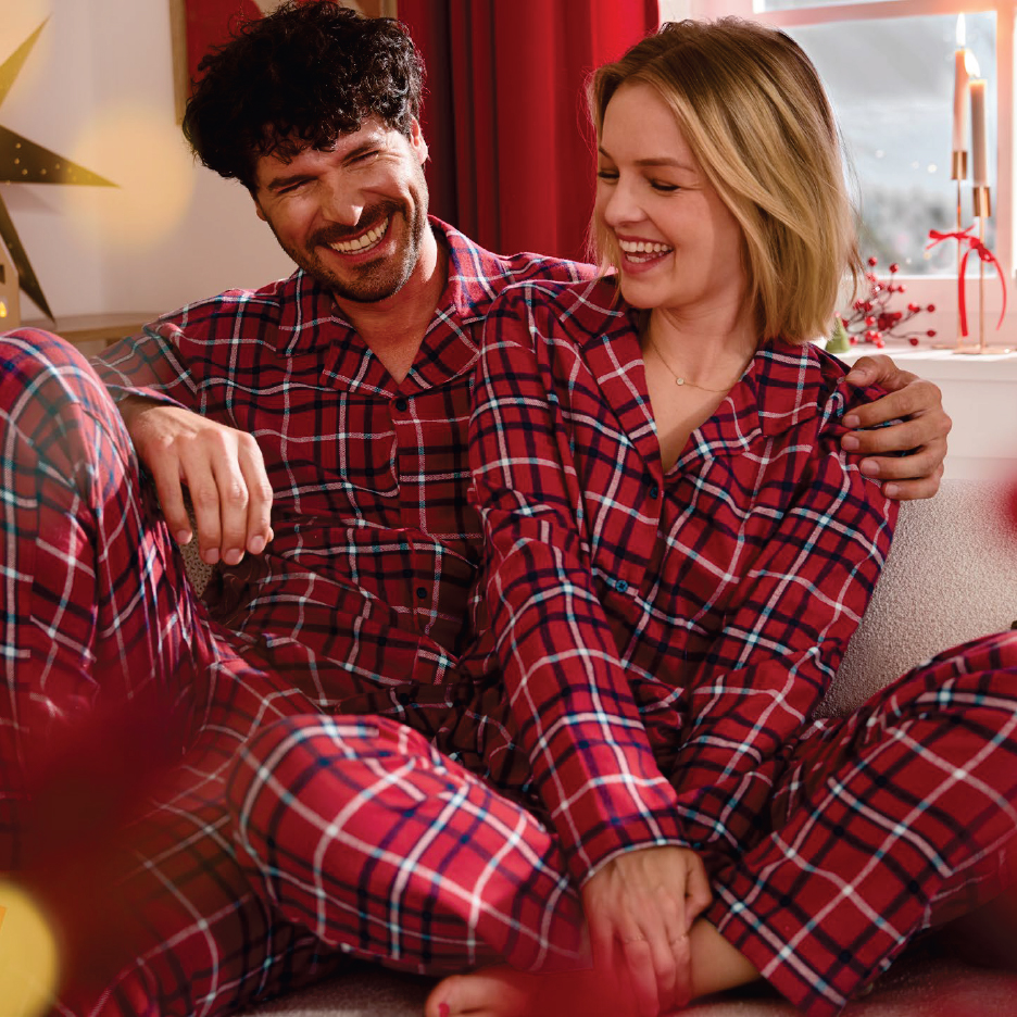 Двойка в червени карирани пижами се смее на диван по време на празниците.