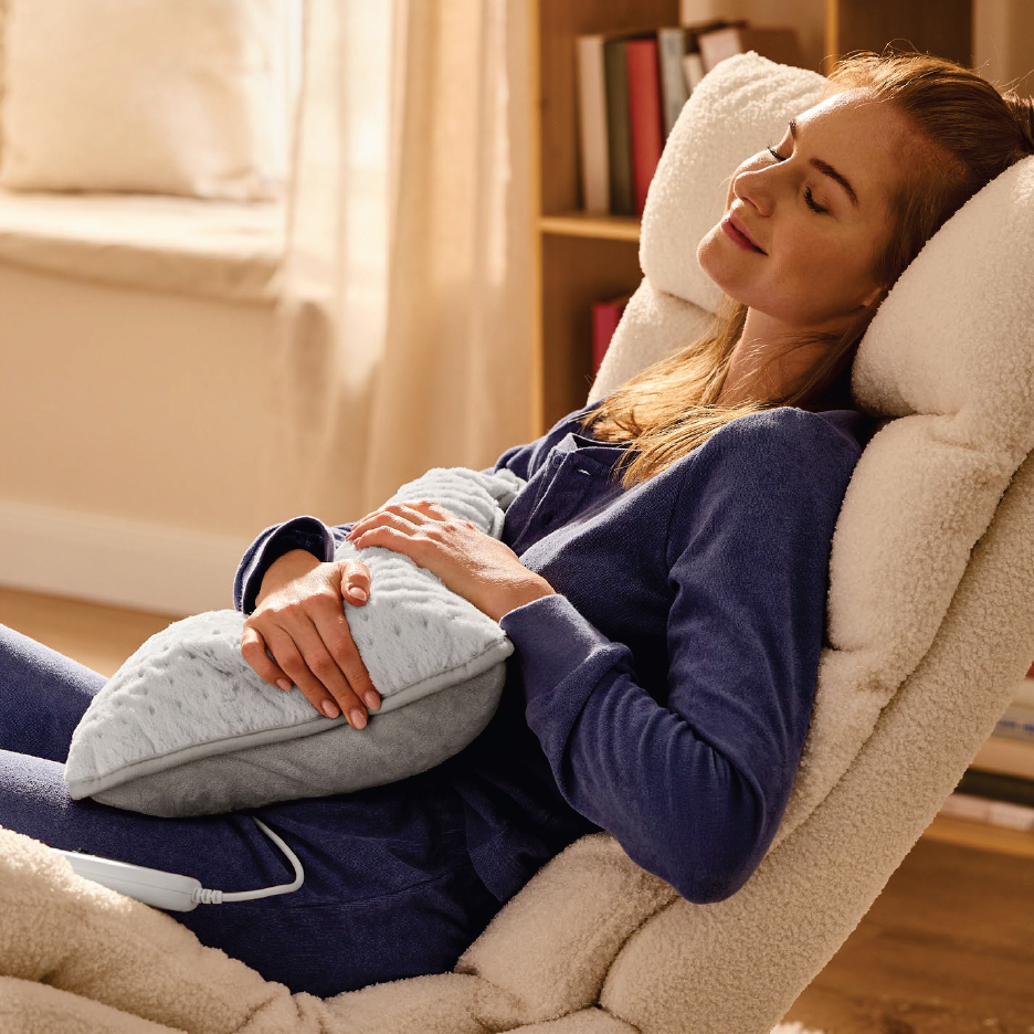 Жена релаксира в кресло, прегърнала електрическа възглавница за гръб.