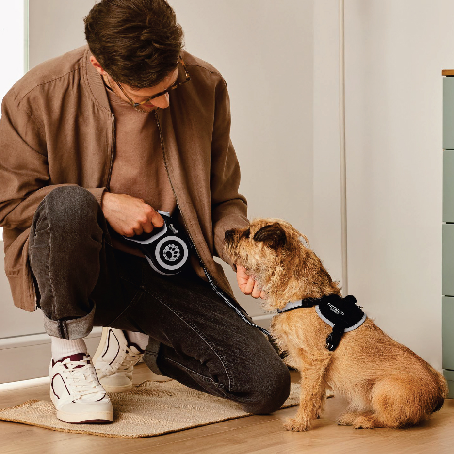 Мъж с кафяво яке и черни дънки, държи черен повод за куче, докато гали куче с кафява козина, носещо черен нагръдник.