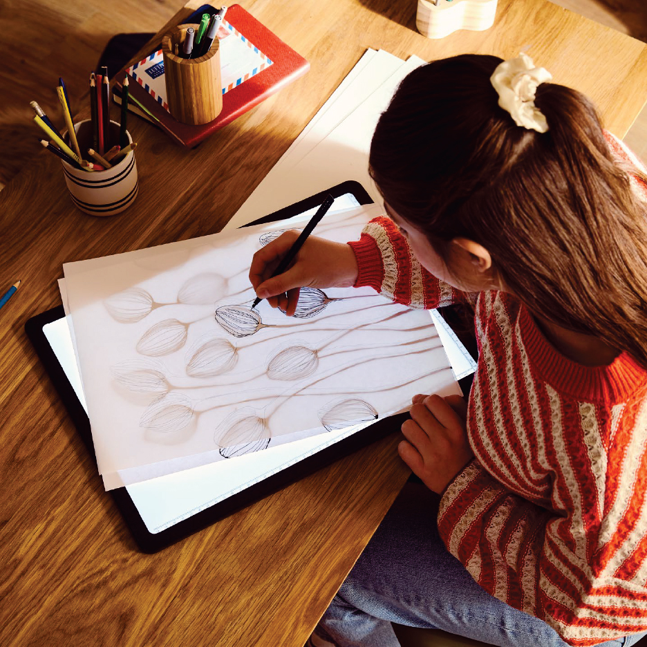 Момиче рисува върху светеща дъска, прекопирайки скица на цветя.