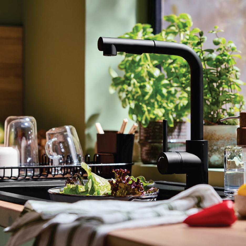 Черна кухненска батерия над мивка със салата, съдове и растения на заден план.