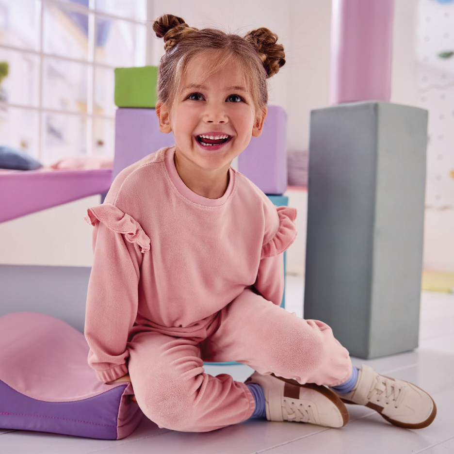 Усмихнато момиченце в розов комплект от суичър и панталон, седнало на пода.