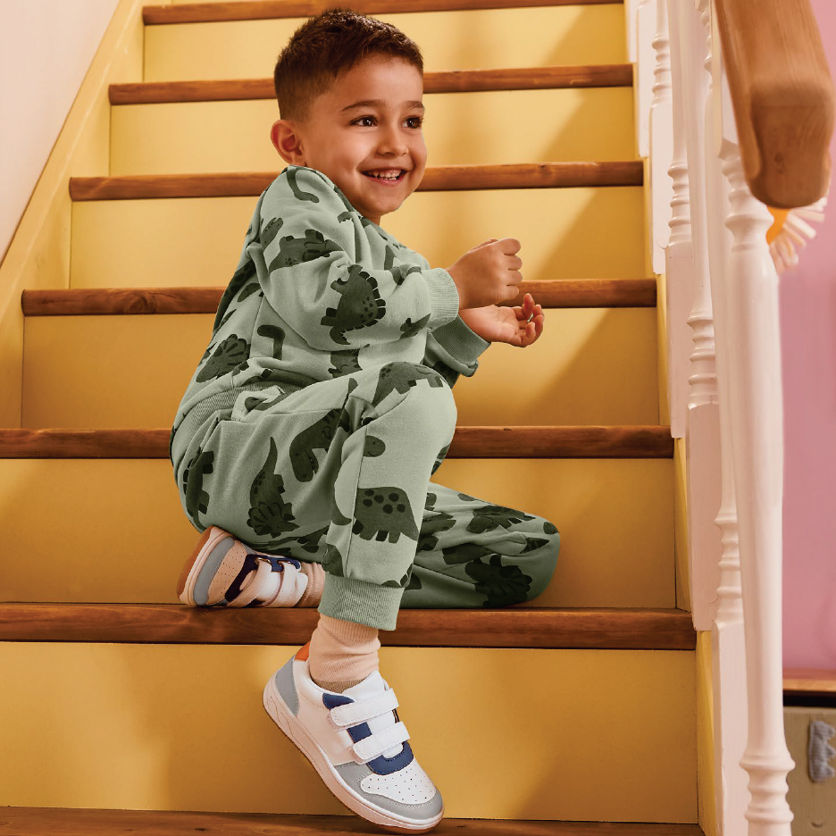 Момче в зелен комплект с динозаври и маратонки седи на стълби.