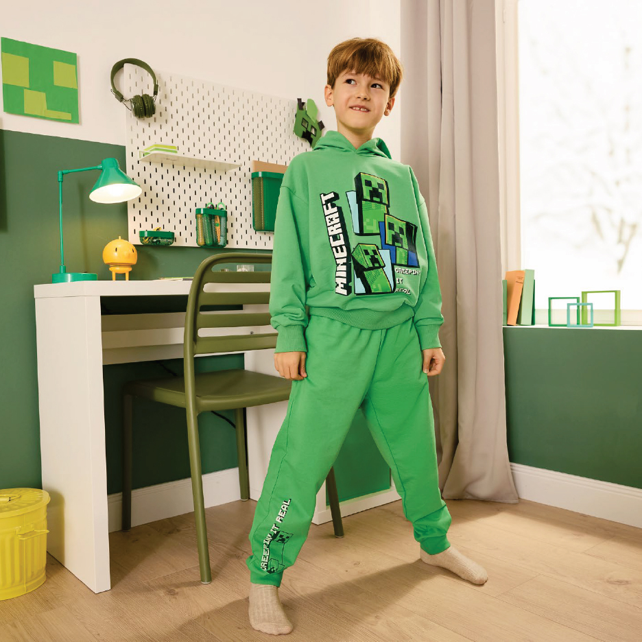 Момче в зелен суичър и долнище с щампа на кубчета, стоящо до бюро.