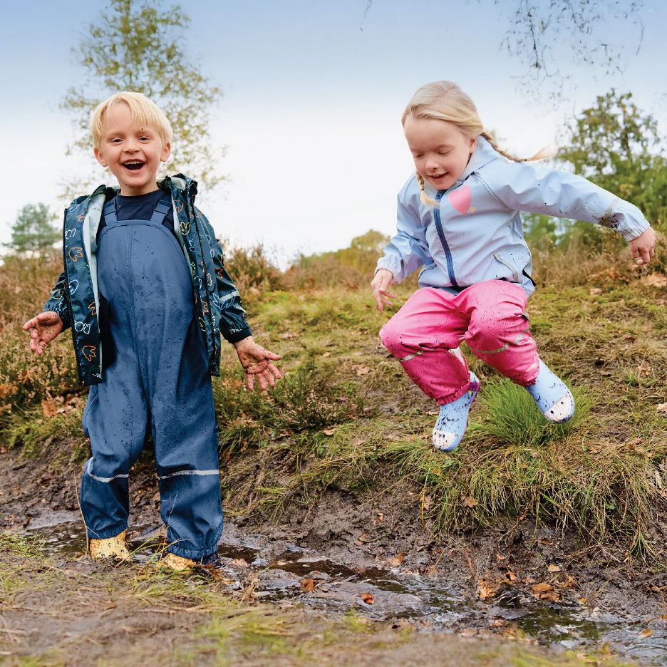 Две деца в непромокаеми гащеризони и якета скачат в кални локви.