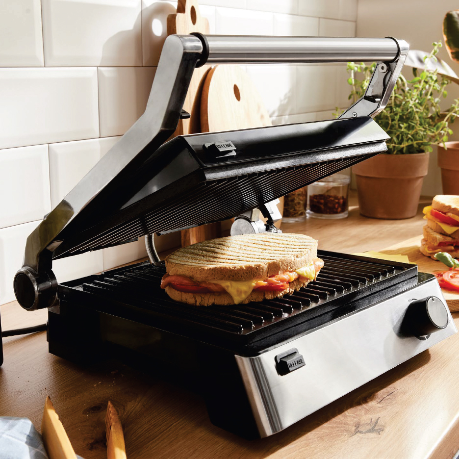 Грил преса с отворен капак, притискаща сандвич със сирене и домати на дървен плот.
