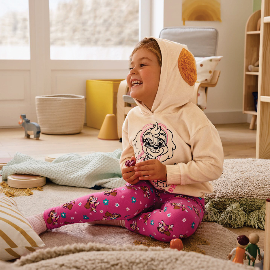 Момиче в розова пижама с Щастливото кученце Скай от Пес патрул, седи на килим в детска стая.