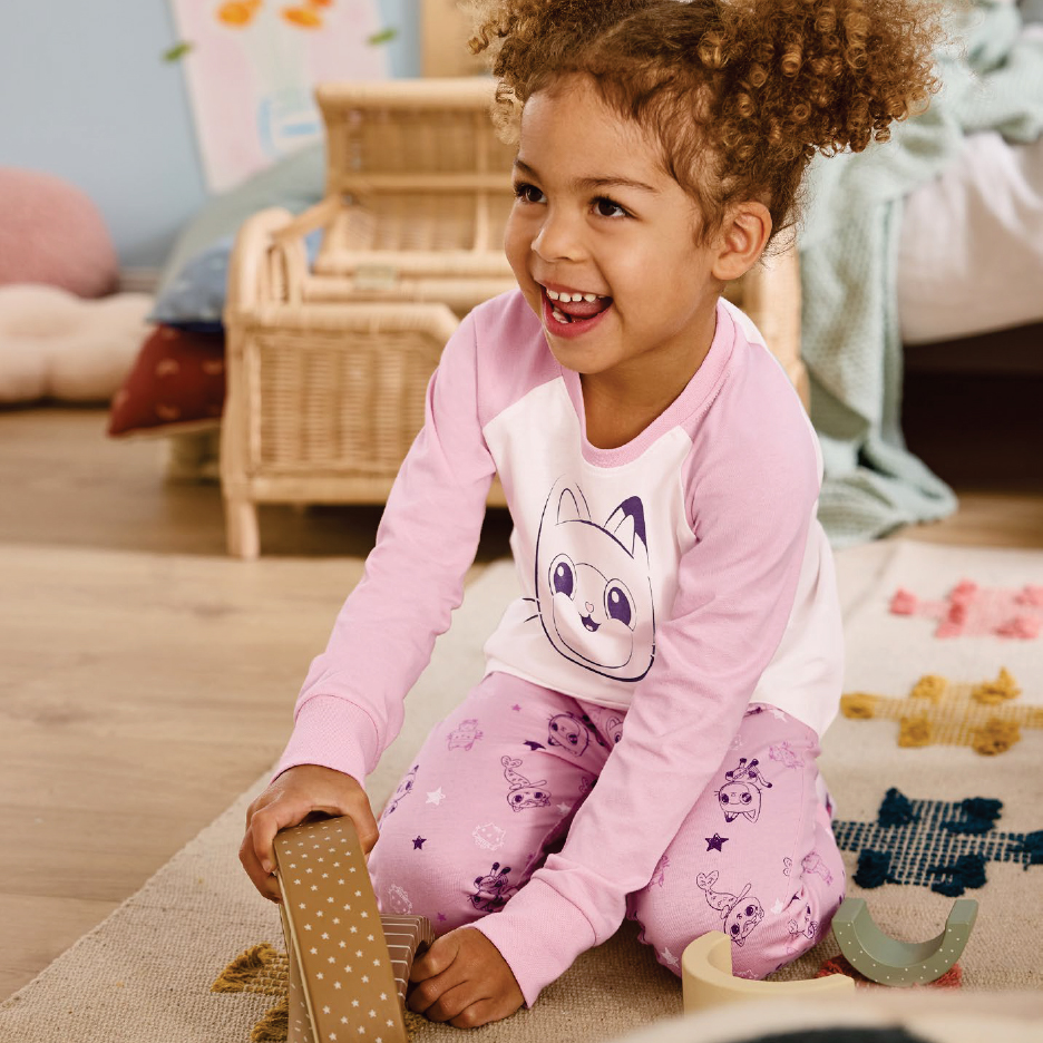 Момиченце в розови пижами с котешки мотиви, играещо на пода.