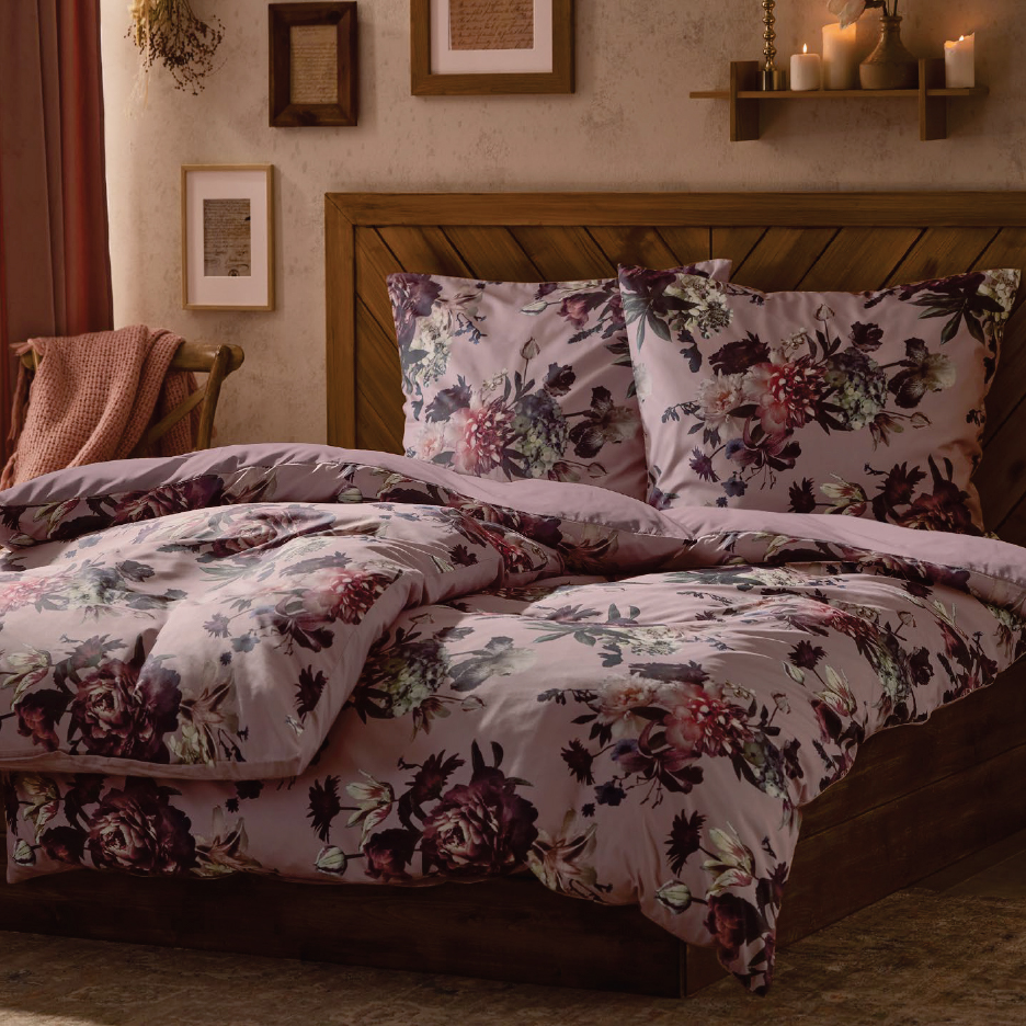 Спалня с флорално спално бельо в лилави нюанси, дървена табла и нощно шкафче.