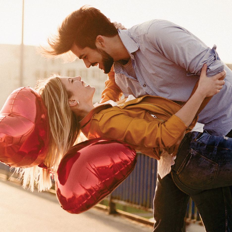 Влюбена двойка с червени балони във формата на сърце, мъжът държи жената в прегръдка.