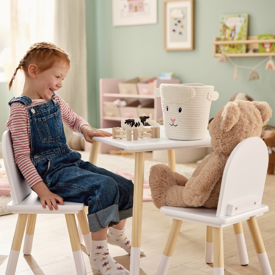 Усмихнато момиченце в гащеризон седи на бяла детска маса с плюшено мече и играчки.