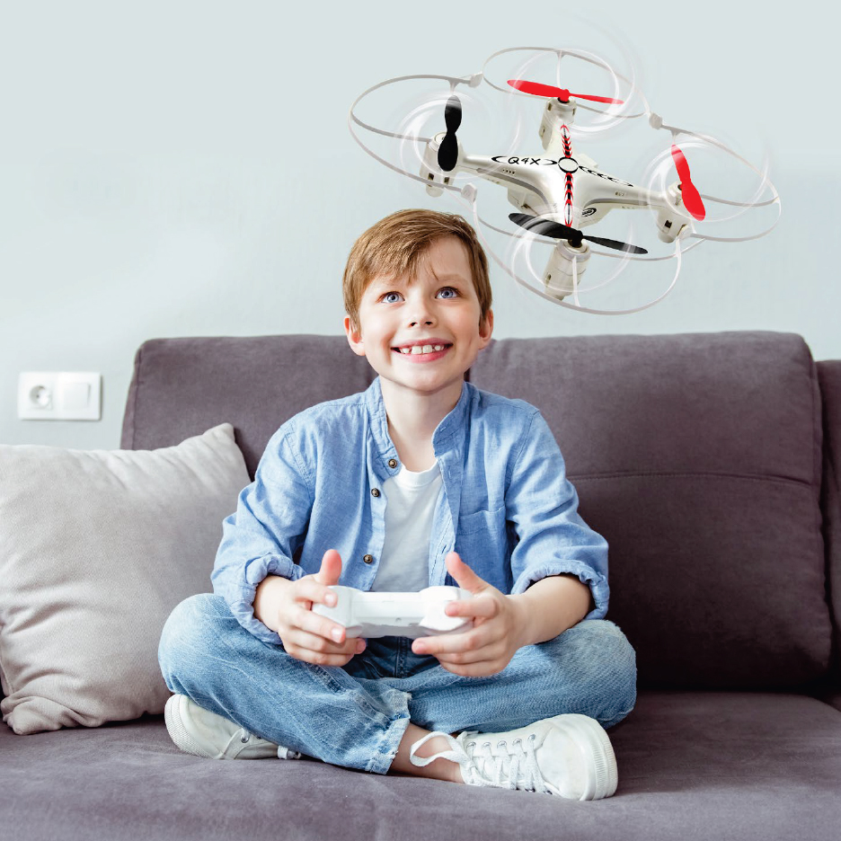 Усмихнато момче играе с бял дрон с червени и черни перки, докато седи на диван.