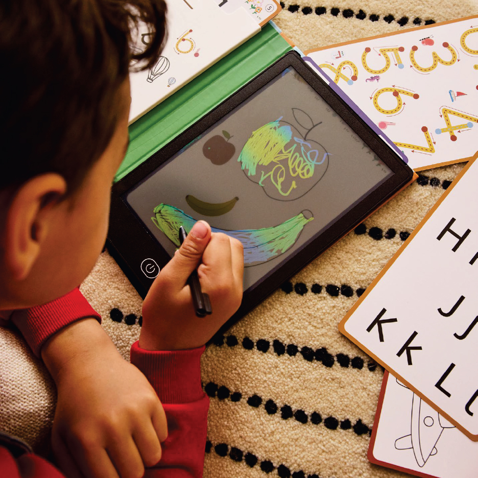 Дете рисува плодове на дигитална дъска, заобиколено от карти с букви и цифри.