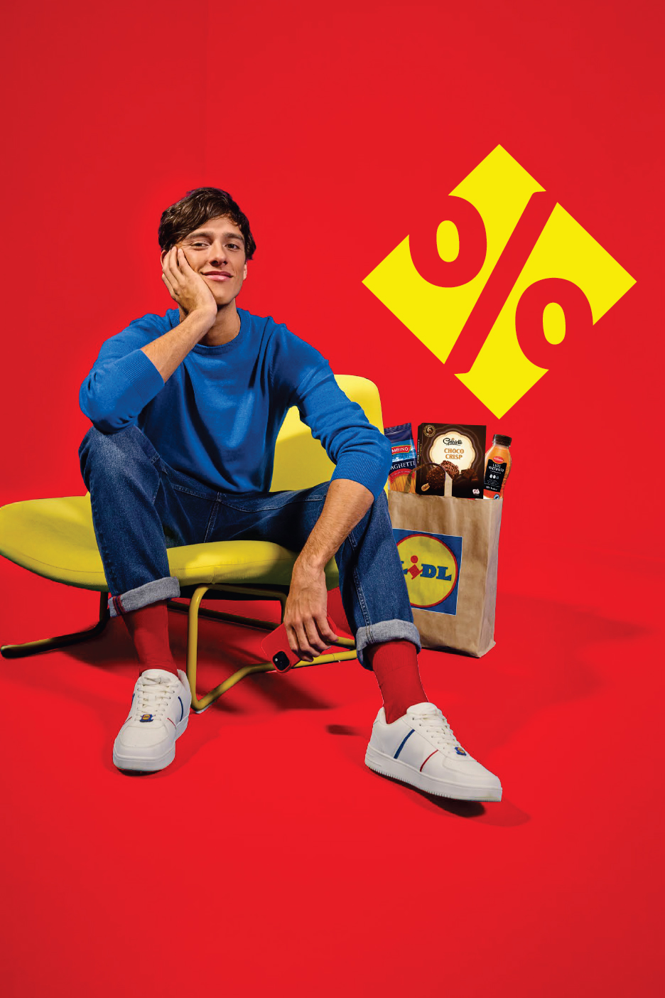Мъж седи до пазарска чанта на Lidl с паста, шоколадов десерт и напитка, на червен фон с жълт знак за процент.