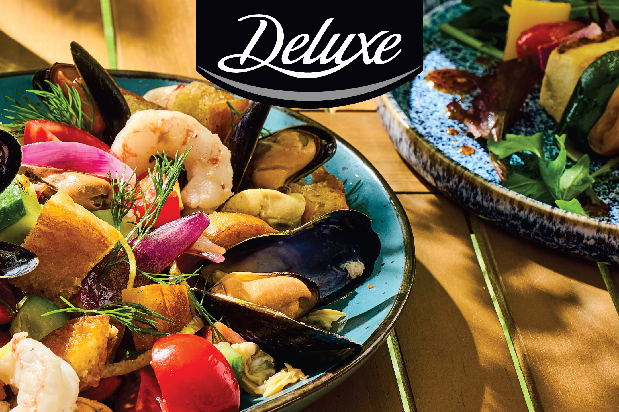 Салата с миди, скариди, домати, лук и крутони в синя купа, с логото „Deluxe“.