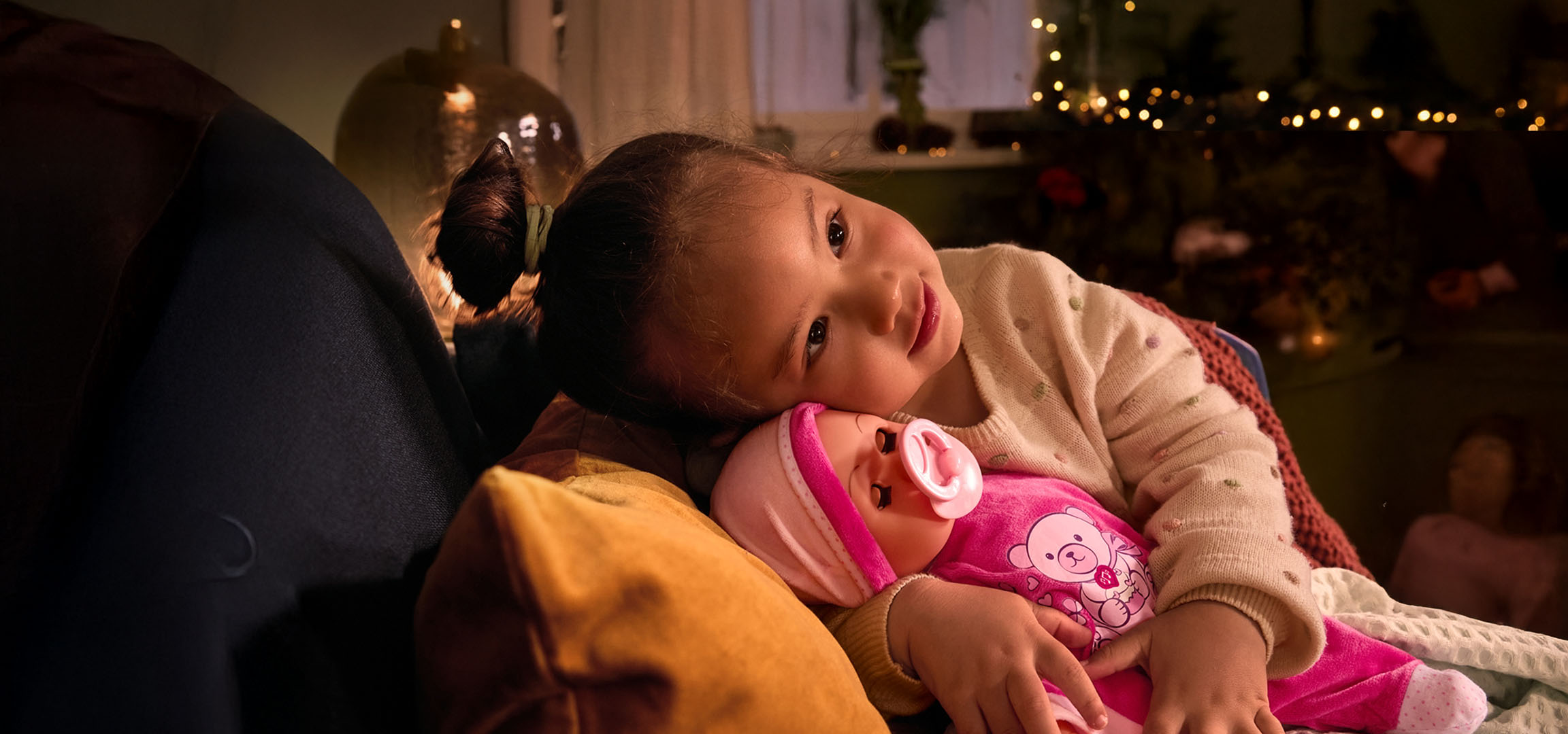 Момиченце с кукла и биберон, удобно настанено на възглавница.