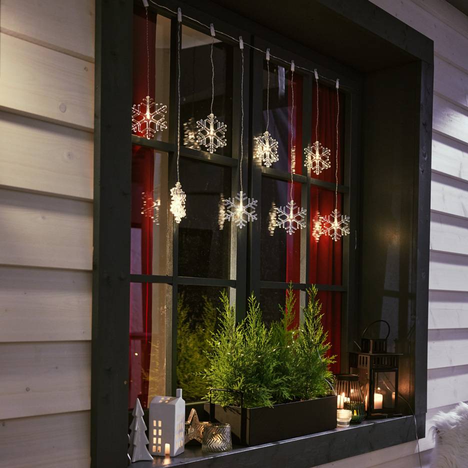 Светещи снежинки висят на прозорец с малки елхички и свещи на перваза.