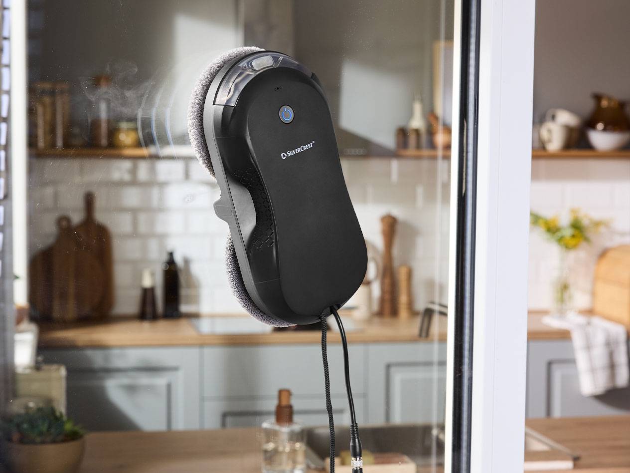 Робот за почистване на прозорци Silvercrest върху стъкло с кухня на заден план.