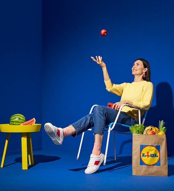 Жена хвърля ябълка, седнала на стол с чанта Lidl, пълна с хранителни стоки.