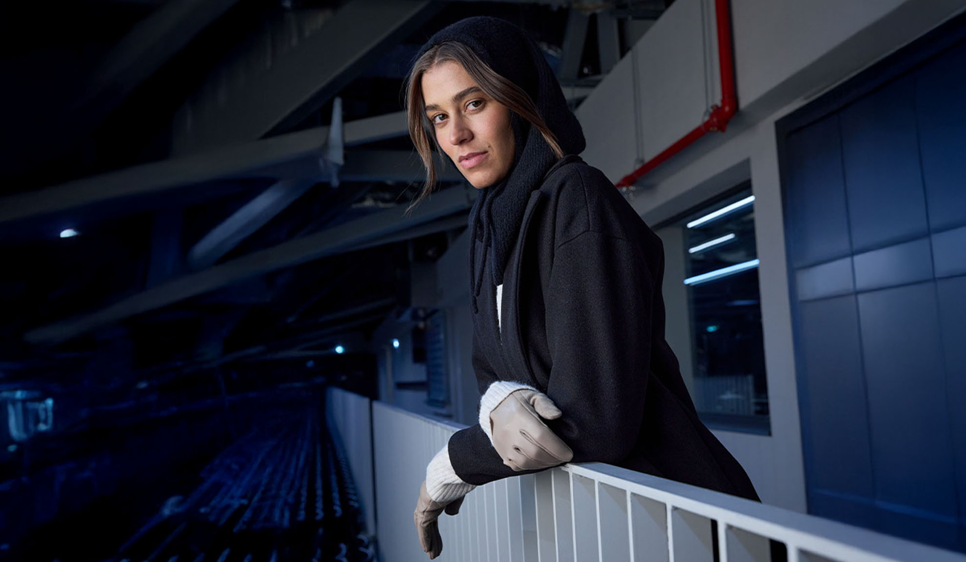 Жена в черно палто и шал с качулка, облегната на парапет в модерна обстановка.