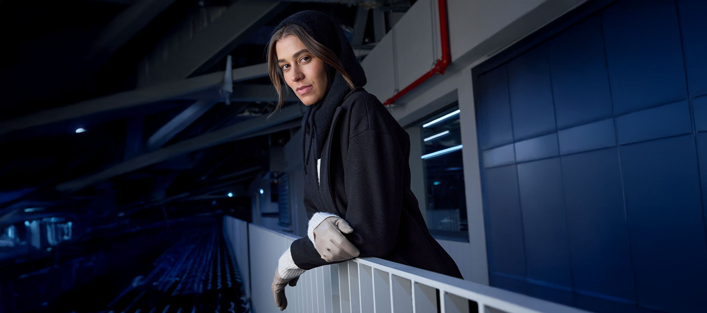 Жена в черно палто и ръкавици, облегната на парапет в градска среда.