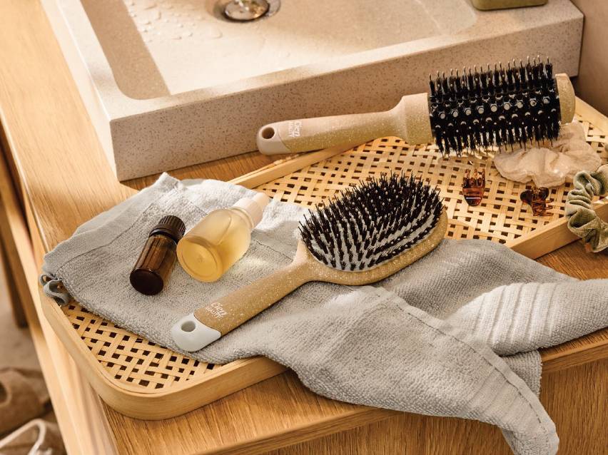 Две четки за коса, една овална и една кръгла, поставени върху сива кърпа до шишенца с масло.