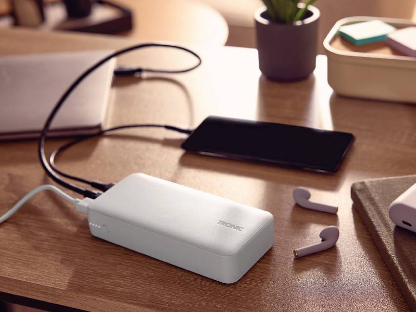 Бяла външна батерия зарежда телефон и лаптоп на дървена маса, до нея са поставени безжични слушалки.